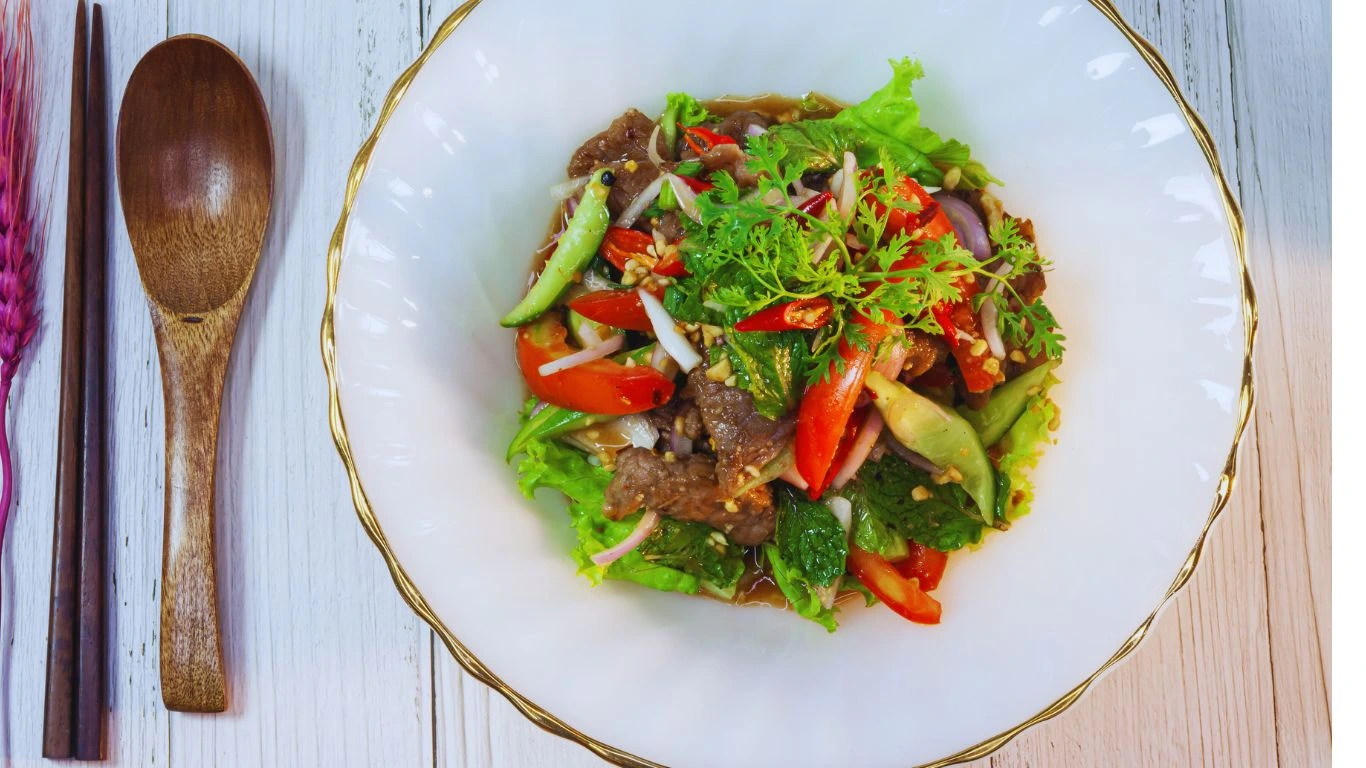 Colorful Thai Beef Salad featuring thinly sliced grilled beef, fresh herbs, cherry tomatoes, cucumber, red onion, and chili, all tossed in a zesty lime-fish sauce dressing, served on a white plate