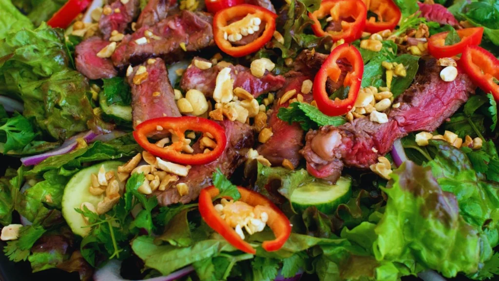 A vibrant Thai Beef Salad with thinly sliced grilled beef, cherry tomatoes, cucumber, red onions, and fresh herbs, served with a spicy lime and fish sauce dressing on a white plate