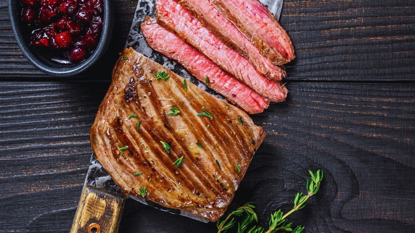 Grilled beef flap steak sliced on a wooden cutting board, served with chimichurri sauce and roasted vegetables.